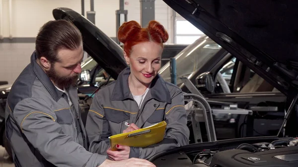 Beautiful female mechanic and her male colleague examining engine of a car