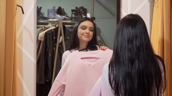 Affascinante giovane donna cercando vestiti di fronte a uno specchio al negozio di abbigliamento — Foto Stock