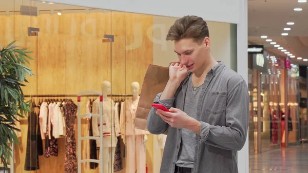 Joven sonriendo, usando un teléfono inteligente mientras hace compras en el centro comercial —  Fotos de Stock