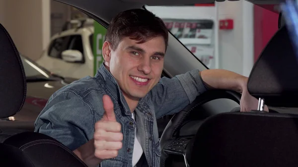 Счастливый красивый мужчина, показывающий большие пальцы вверх, сидящий в новой машине в салоне — стоковое фото