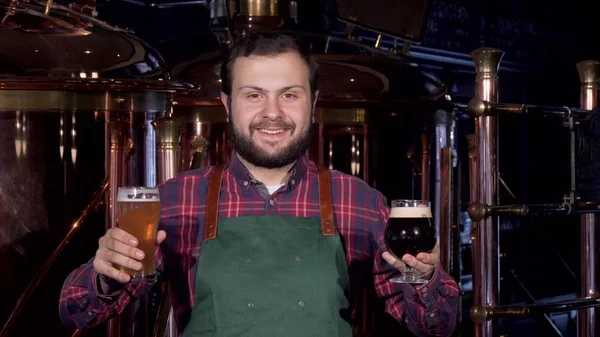 Happy male brewer in apron smiling joyfully holding two glasses of craft beer