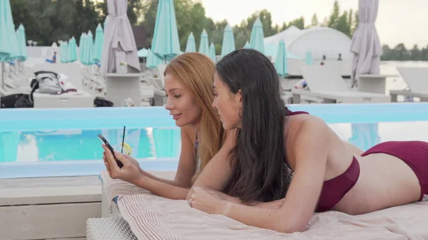 Дві прекрасні жінки використовують смартфон, відпочиваючи біля басейну — стокове фото