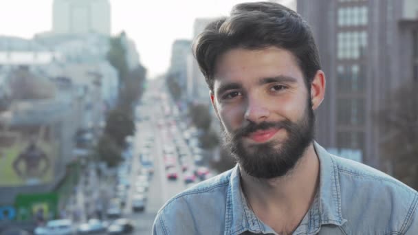 Joven barbudo sonriendo a la cámara con la ciudad en el fondo — Vídeo de stock