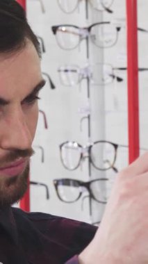 Yakışıklı, sakallı genç bir adamın, modern bir optometri mağazasında canlı bir çift gözlük ile çevrili şık bir gözlüğü dikkatle incelemesi. Moda çerçevelerinin renkli görüntüsü davetkar bir atmosfer yaratıyor.