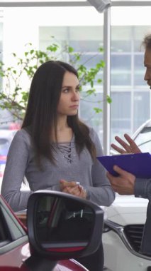 Keskin mavi gömlekli deneyimli, sakallı bir satıcı şık bir modern araba galerisinde genç bir bayanla seçenekleri tutkuyla tartışıyor. Göz kamaştırıcı kırmızı araba, galeri ışıklarının altında parlak bir şekilde parlıyor.