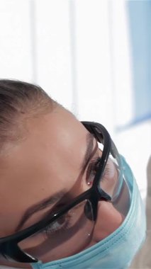 Genç, esmer bir dişçinin yakın plan fotoğrafı. Temiz beyaz bir sağlık üniforması ve açık mavi bir maske takıyor. Parlak bir gülümsemeyle muayeneyi tamamlıyor. Kibarca maskesini çıkarıp profesyonelliği somutlaştırırken dostane tavrı ortaya çıkıyor.