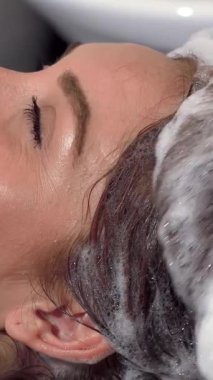 Huzurlu bir sahne, gençleştirici kafa masajından zevk alan güzel bir kadının lüks bir salonda saçlarını köpüklü şampuanla kaplamasını çeker. Kuaförün uzman dokunuşu neşe saçan bir gülümseme ortaya çıkarır.