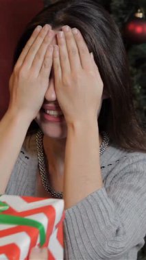 Neşeli genç bir kadın, elleri neşeyle gözlerini kapatıyor, sevimli küçük oğlundan içten bir Noel sürprizi bekliyor. Parıldayan ışıklar ve güzel süslenmiş bir Noel ağacına karşı koyduğunda, heyecanı yayılıyor.