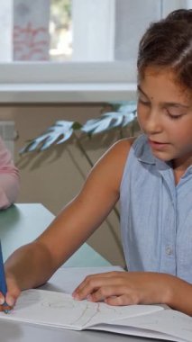 Odaklanmış, uzun saçlı küçük bir kız açık eskiz defterine tutkuyla resim çizerken, gururlu öğretmeni ışıl ışıl bir gülümsemeyle izliyor. Bu iç açıcı sahne, renkli bir sınıfta yaratıcılığın ve cesaretlendirmenin canlı enerjisini yakalıyor.