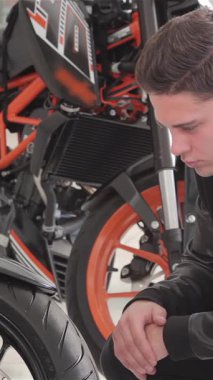 Neşeli beyaz bir adam çarpan siyah bir motosikletin yanında diz çöker ve gülümseyen bir gülümsemeyle baş parmağını kaldırır. Heyecanı, motosikletleri incelerken ışıldıyor gösterişli tasarım ve canlı turuncu aksanlar, macera ve tutku dolu