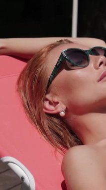 Göz kamaştırıcı kızıl saçlı bir kadın şık bir iskemlede lüks içinde güneşlenerek, parlak bir yüzme havuzunun yanında güneşleniyor. Canlı pembe bir bikini ve şık güneş gözlükleriyle giyinmiş yaz cazibesi ve kaygısız bir mutluluk yayıyor. Mükemmel bir tatilin vücut bulmuş hâli.