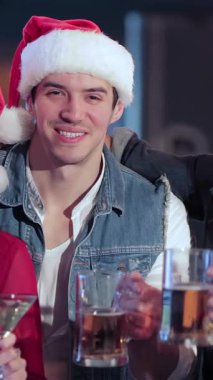 Canlı bir üçlü, iki erkek ve bir kadın, neşeli bir barda kadehlerini kaldırırken neşeli Noel Baba şapkaları giyerler. Atmosfer kahkaha ve bayram neşesiyle uğulduyor. Renkli dekorasyonlar ve sıcaklıkla çevrili.