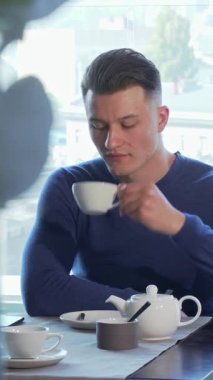 Hoş mavi kazaklı yakışıklı genç bir adam sıcak bir fincan çayın tadını çıkarıyor, canlı bir şehir manzarasına dikkatle pencereden dışarı bakıyor. Bu sakin an zarafet ve rahatlığı simgeler. Kahvaltı toplantısı ya da sessiz tefekkür için idealdir.