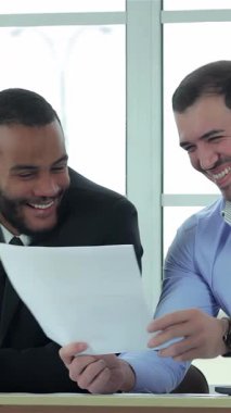 İki kendine güvenen iş adamı, biri parlak siyah takım elbiseli, diğeri açık mavi gömlekli, güneşli bir ofiste belgeleri neşeyle gözden geçiriyorlar. Bulaşıcı gülümsemeleri ve yoldaşlıkları başarılı bir ortaklığı vurguluyor, pozitif enerji yayıyorlar.