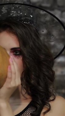 Büyüleyici bir kadın büyücü, kaprisli bir cadı şapkasıyla süslenmiş, şakacı bir şekilde yüzünü sırıtan bir balkabağının arkasına gizliyor, dramatik bir siyah tuğla duvara dayanıyor. Bu Cadılar Bayramı temalı sahne bir gizem ve sihir havası yayıyor.