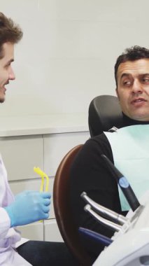 Kendini işine adamış genç bir dişçi, el değmemiş beyaz bir ceket giymiş, neşeli bir şekilde gülümseyen erkek hastasına ayna uzatıyor, o da gösterişli, modern bir dişçi koltuğunda rahat bir şekilde oturuyor. Parlak, steril klinik bir güven ortamı yayıyor.