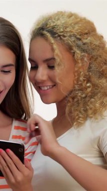 İki kadın, biri kıvırcık kıvırcık sarı saçlı, diğeri uzun kahverengi saçlı, bir tabletle hareketli, yüzleri heyecanla ışıldıyor. Yumuşak beyaz arka plan onların neşeli ifadelerini ve günlük kıyafetlerini geliştirir.