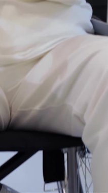 Üst düzey bir beyaz kadın parlak, modern bir rehabilitasyon odasında etrafındakileri yansıtırken düşünceli bakışları aşağıya doğru çevrilmiş parlak bir tekerlekli sandalyede oturuyor. Yumuşak, ortam aydınlatması onun nazik özelliklerini vurgulayarak, bir huzur yaratıyor.