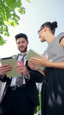 Kendinden emin bir işadamı ve profesyonel bir iş kadını, canlı yeşil yapraklardan oluşan bir gölgeliğin altında sıcak güneş ışığının tadını çıkararak tabletlerinde fikirlerini coşkuyla paylaşıyorlar. Onların ışıltılı gülümsemeleri ve çekici ifadeleri başarılı bir şekilde yansıyor.