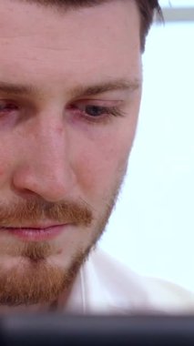 Genç, çekici bir beyaz erkek yönetici öne eğiliyor, delici mavi gözleri bilgisayar ekranına kilitlenmiş, modern ofis pencerelerinden yansıyan parlak doğal ışıkla aydınlatılıyor. Şık beyaz gömleği ve bakımlı sakalı yansıtıyor.