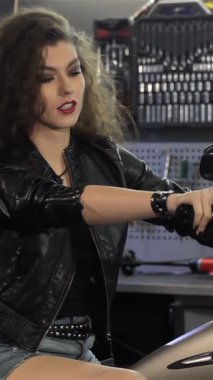 Kıvırcık saçlı ve şık siyah deri ceketli neşeli genç bir kadın, canlı bir atölyede parlak motorsikletine tünediği sırada kendinden emin bir şekilde onay verirken heyecan saçıyor. Bu çekici beyaz motorsikletli,