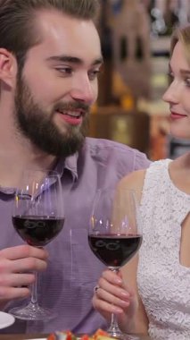 Neşeli genç bir çift sıcak bir restoranın ışıltısında güneşlenen koyu kırmızı şarap kadehlerini paylaşır. Işıl ışıl gülümsemeleri ve samimi bakışları güzel tabaklar ve yumuşak tabaklarla güçlendirilmiş büyülü bir atmosfer yaratıyor.