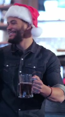 Neşeli bir barda, Noel Baba şapkası takan iki arkadaş bayram ruhunu somutlaştırmak için buzlu bira bardaklarını kaldırır. Arka plandaki neşeli barmen ışıltıları neşe, neşe ve neşe dolu neşeli atmosferi yükseltiyor.