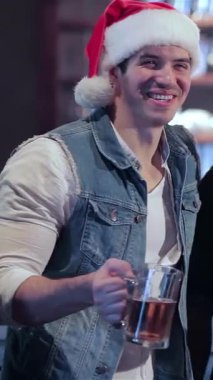 Şenlikli süslemelerle süslenmiş hareketli bir barda Noel Baba şapkalı iki arkadaş bayram ruhunu somutlaştıran coşkulu bir tostla buzlu bira bardaklarını kaldırıyor. Arka planda neşeli barmen ışıltıları ile dolu canlı atmosfer