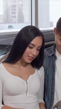 Neşeli bir çift sıcak bir kucaklaşmayı paylaşıyor, parlak yeni araba anahtarlarını lüks araçlarla dolu, modern bir galeride gururla sergiliyorlar. Güzel kadın heyecanla ilk otomobillerini satın almalarının kilometre taşını kutluyor.