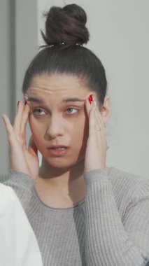 Yüzü acıyla oyulmuş genç bir kadın, doktoruna danışır, şık, modern bir klinikte, kafasını hüsran içinde tutarak. Parlak ve klinik ortam, migren semptomlarının aciliyetini vurgulayan ciddi ifadesiyle çelişiyor.