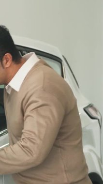 Siyah saçlı, kendine güvenen bir adam, parlak beyaz bir arabanın açık camına doğru eğiliyor, lüks iç mekanını keskin bir ilgiyle inceliyor. Nazikçe dikiz aynasına dokunuyor, sonra sıcak, davetkar bir gülümsemeyle kameraya dönüyor, ışıltılı bir cazibeyle ve