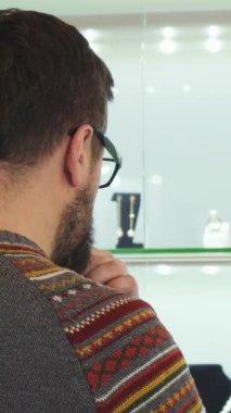 Sade, desenli süveterli sakallı bir adam hareketli bir alışveriş merkezinde durup göz kamaştırıcı bir mücevher sergisini dikkatle inceliyor. Düşünceli tavrı, yürekten bir hediye seçmenin heyecanını yakalıyor. Havada romantizm ve zerafet yayıyor.