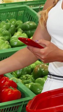 Şık beyaz elbiseli şık bir kadın, şık bir süpermarkette yemyeşil sebzelerden aldığı parlak kırmızı biberleri dikkatle seçerken akıllı telefonuna hayran kalıyor. Bu dinamik sahne modern dünyanın heyecanını güzel bir şekilde birleştiriyor.
