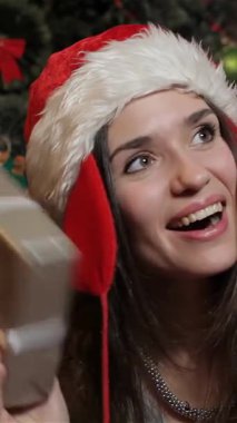 Neşeli esmer bir kadın, neşeli bir Noel Baba şapkası takıyor, güzel paketlenmiş bir Noel hediye kutusunu neşeyle sallıyor, yüzü saf zevk saçıyor. Işıl ışıl süslerle ve parıldayan ışıklarla süslenmiş ışıl ışıl bir Noel ağacıyla çevrili.