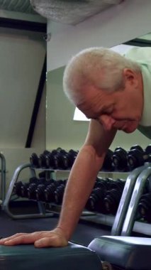 Gücünü ve kararlılığını sergileyen yaşlı beyaz bir adam dizini şık bir spor salonunda dinlendirirken dambılı kaldırır. Destekleyici genç bir antrenör, bu canlı fitness alanında motive edici bir atmosferi teşvik etmek için yakında duruyor.