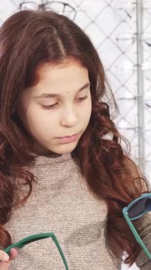 Kahverengi bukleli tatlı bir kız canlı bir gözlük dükkanında elinde iki tane şık güneş gözlüğü ile duruyor. Yüzü kafa karışıklığı ve merakı yansıtıyor. Renkli gözlüklerden oluşan bir kaleydoskop ile çevrelenmiş,