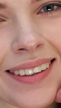 Güzel beyaz bir kadının büyüleyici yakın çekimi kusursuz makyajıyla kendine güveni yayıyor, nazikçe yüzüne dokunuyor. Işıl ışıl cildi el değmemiş beyaz bir zemin üzerinde parıldıyor, büyüleyici gözlerini ve tatlı gülümsemesini aydınlatıyor.