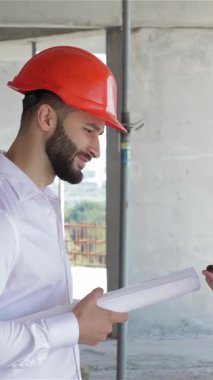 İki genç müteahhit, biri parlak turuncu bir başlık takıyor, diğeri ise klasik beyaz bir şapka takıyor ve hareketli bir inşaat sahasında lüks bir dijital tabletin planlarını tartışıyorlar. Sakallı adam, sarılı bir planı olan, paylaşıyor.