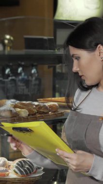 A dedicated young female baker, dressed in a stylish apron, carefully checks stock while jotting down notes in her vibrant yellow clipboard. Surrounded by an enticing display of freshly baked pastries, her passion for her craft radiates, creating a