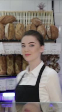 A joyful young woman baker radiates happiness as she warmly welcomes new customers into her charming bakery, surrounded by an enticing display of freshly baked breads and pastries. The delightful aroma of golden loaves and sweet confections fills the