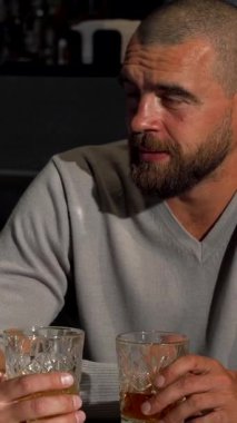 A vibrant scene unfolds as mature male friends share hearty laughter over whiskey glasses in a chic bar setting. The cheerful bearded man, dressed in a stylish sweater, radiates warmth and camaraderie, creating an inviting atmosphere filled with joy
