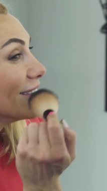 A close-up of a stunning mature woman joyfully applying blush with a soft brush, her radiant smile exuding confidence and beauty. The bright salon ambiance, filled with elegant beauty tools, creates a chic atmosphere that invites viewers into her