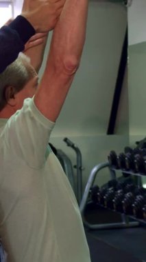 A determined Caucasian senior man lifts a dumbbell with precision, showcasing his strength and dedication in a vibrant gym setting. A supportive young bearded trainer stands behind him, ensuring proper form and motivation, creating an inspiring