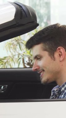 A cheerful young man with a radiant smile sits confidently in a sleek convertible, exuding excitement and style. Surrounded by the modern elegance of the dealership, he grips the steering wheel, embodying the thrill of luxury and new adventures