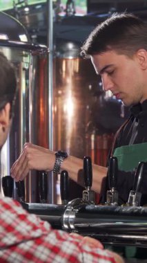 A charismatic male bartender, clad in a sleek black shirt and vibrant green apron, skillfully pours a frothy craft beer into a glass, delighting his eager customer. The lively atmosphere of the bustling pub, with gleaming copper brewing tanks in the