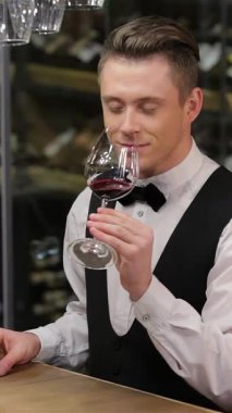 A confident sommelier in a sleek waistcoat and bow tie savors a glass of deep red wine, inhaling its rich aromas with a look of delight. Set against a backdrop of an elegant wine shelf, this scene radiates sophistication and expertise, inviting wine