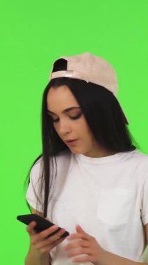 Uzun siyah saçlı ve modaya uygun şapkalı hayat dolu genç bir kadın akıllı telefonuyla canlı yeşil bir krom arka planda selfie çekiyor. Neşeli gülüşü ve neşeli tavırları modern sosyal medyanın özünü yansıtıyor ve izleyicileri davet ediyor.