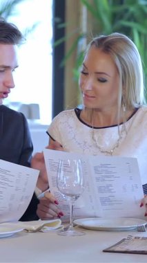 Çok güzel süslenmiş bir restoranda, hoş bir çift zarif bir sette oturur, yemek menüsünü seçmeye dalmış bir şekilde. Ortam aydınlatmasının yumuşak parıltısı ve çiçeklerin narin düzenlemeleri onların neşeli ifadelerini artırıyor.