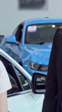 Hevesli bir bayan müşteri, ışıldayan araçlarla çevrili modern bir galeride kendinden emin bir satıcıdan neşeyle araba anahtarı alır. Bu dinamik sahne, yeni sahipliğin heyecanını özetliyor. Başarılı olmanın sevincini gösteriyor.