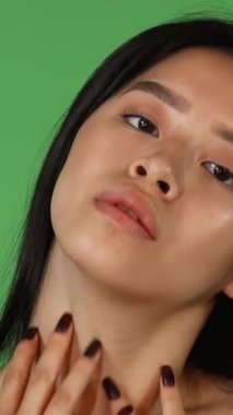 Nefes kesen Asyalı bir kadın canlı yeşil bir krom arka planda zarifçe poz verir narin parmakları yavaşça parlak yüzünü izler. Bu büyüleyici portre kendine güven ve güzellik yayıyor, onun gençlik cazibesini ve doğal cazibesini yakalıyor.